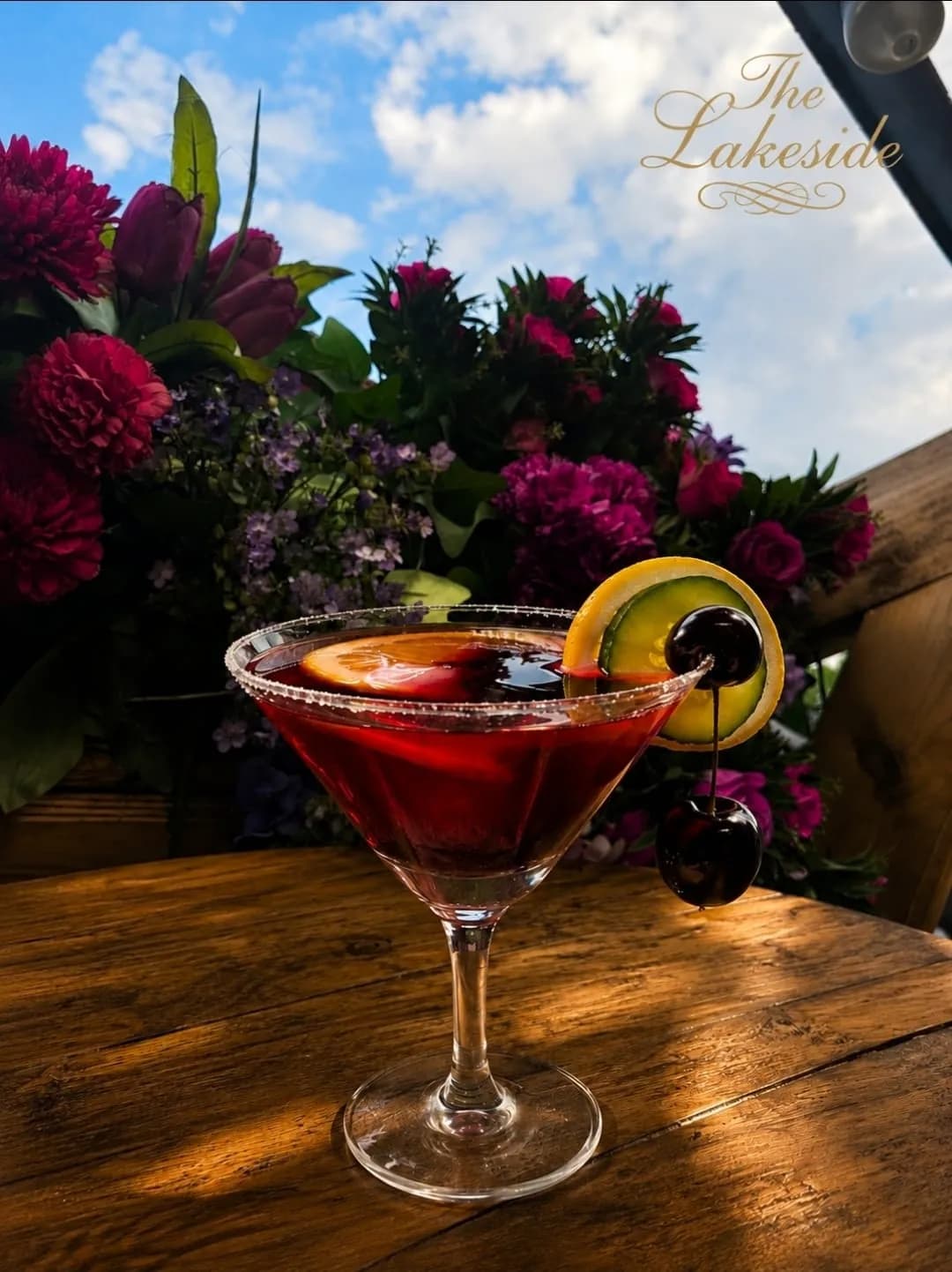 A deep red cocktail in a sugar rimmed martini glass with lemon, cucumber and cherries, in front of pink and purple flowers