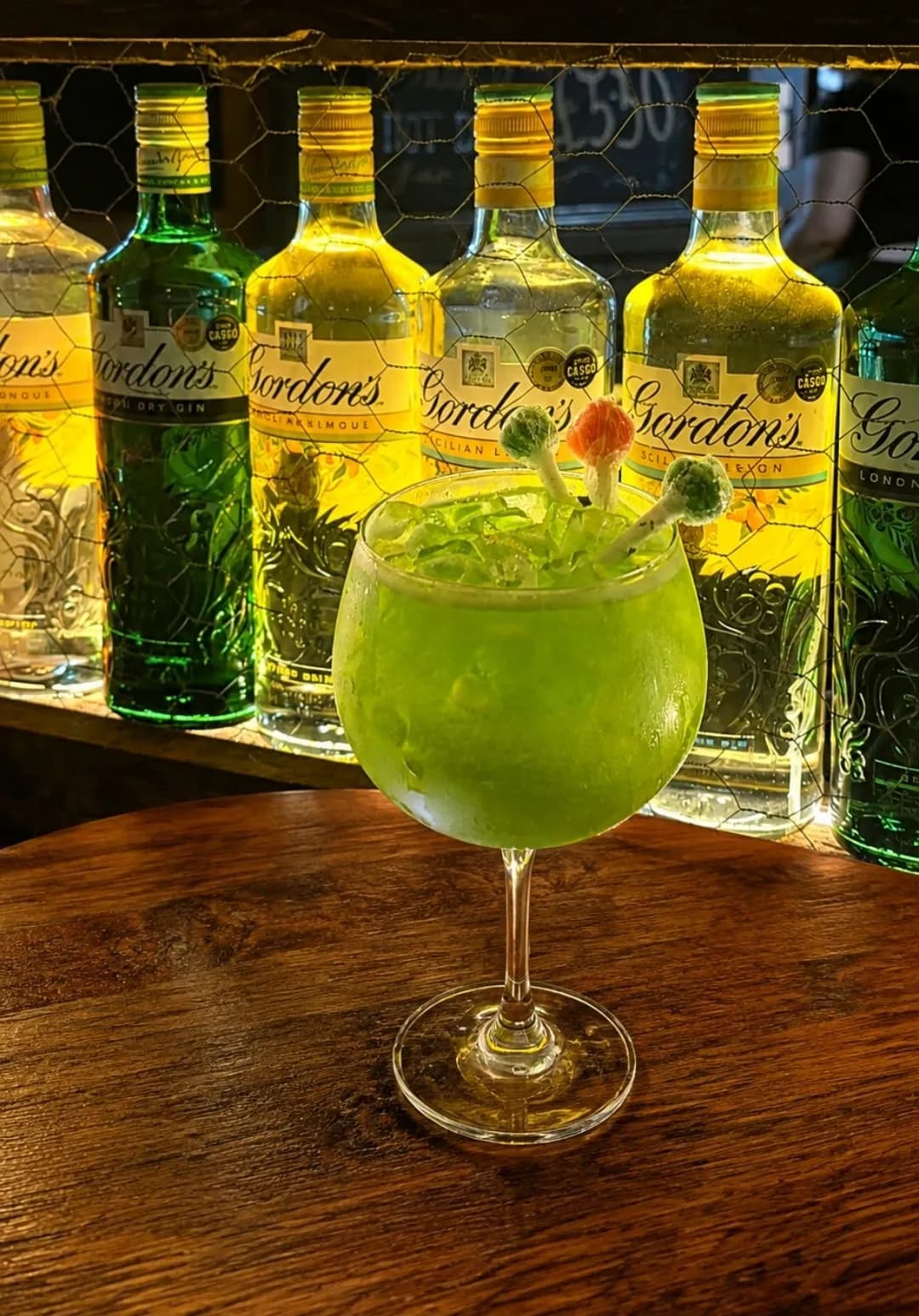 A bright green gin cocktail in a balloon glass, topped with sweets, in front of a row of backlit Gordon's gin bottles