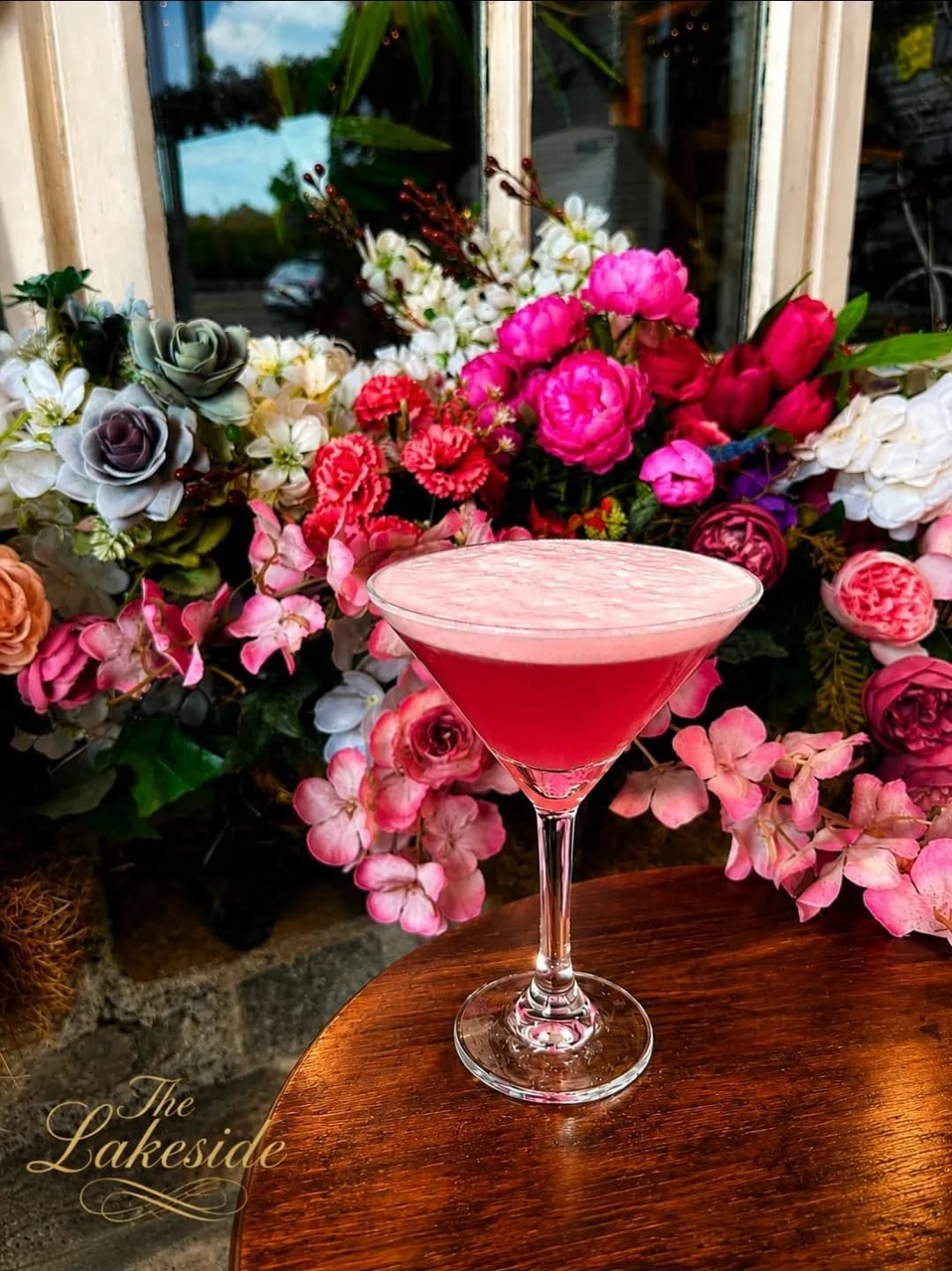 A frothy pink cocktail in a martini glass in front of a wall of pink and white flowers